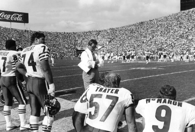 May include: Black and white photo of a football coach and players on the field. The coach is wearing a white shirt and tie, and the players are wearing jerseys with numbers. The photo was taken at a football game.