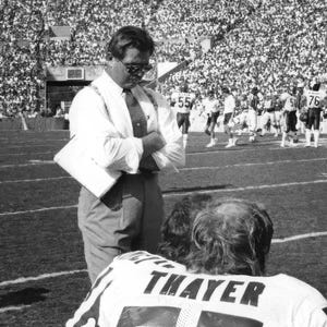 May include: A black and white photo of a man in a suit and tie standing on a football field. He is talking to a young man wearing a jersey with the name "Thayer" on the back.