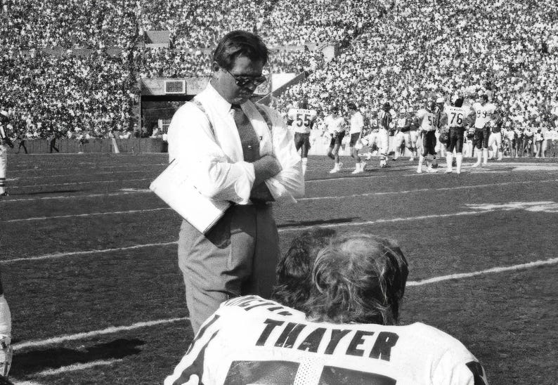 May include: A black and white photo of a man in a suit and tie standing on a football field. He is talking to a young man wearing a jersey with the name "Thayer" on the back.