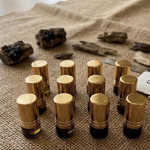 May include: An assortment of small, gold-capped perfume bottles arranged on a textured, tan surface. The bottles are cylindrical with a dark liquid visible inside. The word "OUD" is written on a small white card.