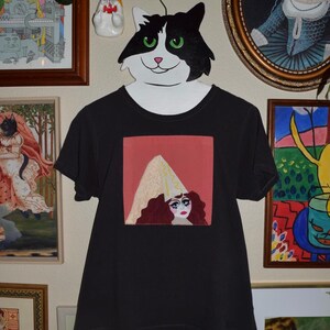 May include: Black t-shirt with a square graphic featuring a woman with red hair and a white hat. The shirt is hanging on a cat-shaped hanger. Various framed artworks and a ceramic cat figurine are in the background.