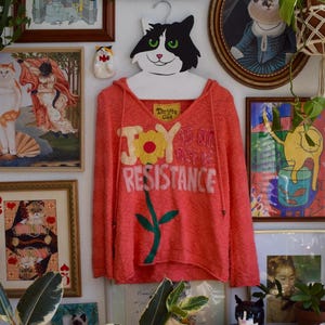 Op de afbeelding: Een koraalkleurige hoodie met de witte tekst "JOY is an act of RESISTANCE", een gele bloem en een groene stengel. De hoodie hangt aan een katvormige hanger, omringd door kunst en decoraties met katten als thema.
