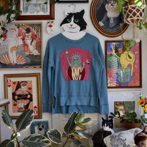 May include: A blue sweatshirt with a graphic of the Statue of Liberty holding a torch and a cigar, with "NYC" text. The sweatshirt hangs on a cat-shaped hanger, surrounded by framed cat-themed art and plants.