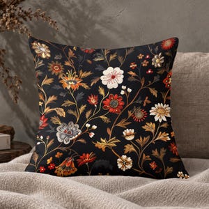 May include: A square throw cushion featuring a black background with a dense, intricate pattern of stylised flowers and leaves in shades of red, orange, gold, white, and grey. The botanical design covers the entire front surface of the cushion.