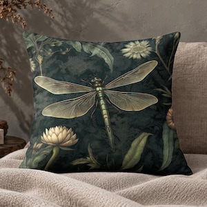 May include: A square throw pillow featuring a dark forest green background with a central dragonfly illustration. The design includes detailed botanical elements such as pale yellow lotus flowers and lush green foliage surrounding the insect.