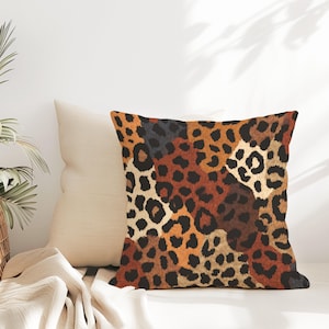 May include: Square decorative pillow with a leopard print pattern in shades of brown, black, and tan. The pillow is placed next to a solid cream-colored pillow. The pillows are on a white surface.