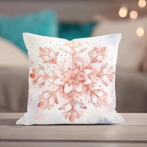 May include: A square decorative pillow with a watercolor snowflake design. The snowflake is in shades of peach and pink, set against a white and light blue background. The pillow is placed on a wooden surface.