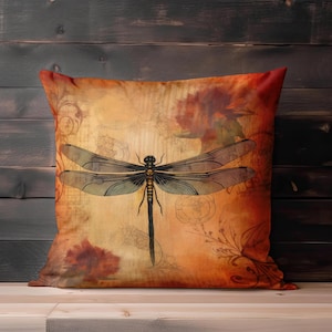 May include: Square decorative pillow with a dragonfly design. The pillow features a vintage-style illustration of a dragonfly in black and gold against a warm, orange and brown background with floral accents.