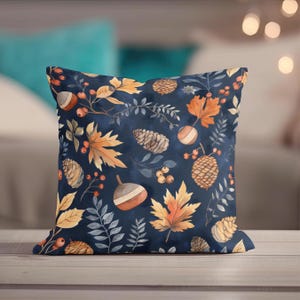 May include: Square cushion featuring a dark blue backdrop adorned with a seasonal autumn design. The pattern showcases illustrations of orange and yellow leaves, acorns, pine cones, and berries. The cushion is likely made of a soft fabric.