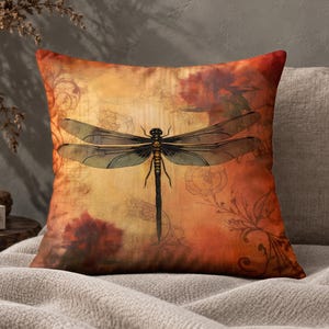 May include: A square throw pillow featuring a central dragonfly illustration with translucent wings. The background displays a warm gradient of orange, gold, and deep red tones, accented with faint, intricate line drawings of floral and scroll patterns.