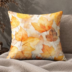 May include: A square throw cushion featuring a watercolour-style pattern of autumn leaves in shades of yellow, orange, and brown against a cream-coloured background. The design displays overlapping maple-like foliage with soft, blended edges.