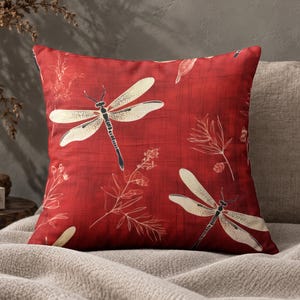 May include: A square throw cushion featuring a deep red background with a subtle cross-hatch texture. The design includes cream-coloured dragonfly illustrations and delicate line-drawn botanical patterns scattered across the front surface.