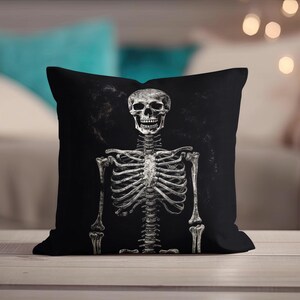 May include: Black decorative throw pillow featuring a detailed, grayscale illustration of a human skeleton. The skeleton is centered on the pillow, with the skull and rib cage clearly visible against a dark background. The pillow is likely made of a soft fabric.