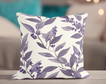 May include: Square cushion featuring a cream-coloured background adorned with a repeating pattern of lavender-hued leaves and floral sprigs. The cushion rests on a wooden surface, with a blurred backdrop.