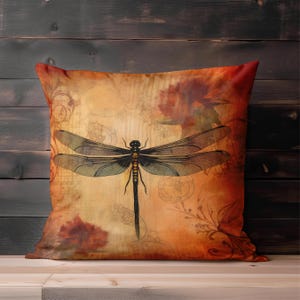May include: Square throw pillow with a dragonfly design. The pillow features a vintage-style illustration of a dragonfly in black and gold against a warm, orange and tan background with floral accents.
