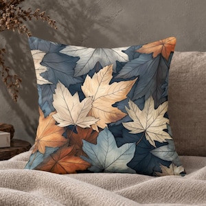 May include: A square throw pillow featuring a dense, overlapping pattern of maple leaves in shades of muted blue, cream, beige, and burnt orange. The design covers the entire front surface with detailed leaf veins and varied autumn-inspired tones.