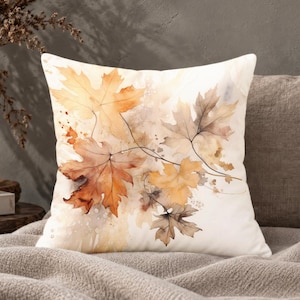 May include: A square throw pillow featuring a watercolor illustration of autumn maple leaves in shades of orange, gold, and brown against a cream-colored background. The design includes delicate leaf veins and soft paint splatters.