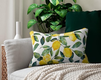 May include: A rectangular decorative pillow with a white background featuring a watercolor pattern of yellow lemons and green leaves. The pillow is placed on a gray sofa with a cream-colored blanket and a green pillow.