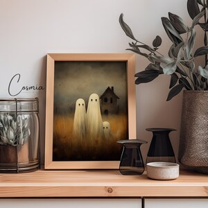 May include: A framed print featuring a spooky scene with three ghost figures in a field, a house in the background, and the text "Cosmia". The artwork is in a wooden frame, displayed on a wooden shelf with decorative items.