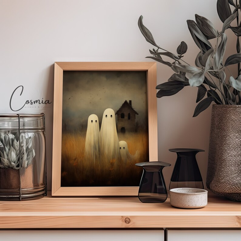 May include: A framed print featuring a spooky scene with three ghost figures in a field, a house in the background, and the text "Cosmia". The artwork is in a wooden frame, displayed on a wooden shelf with decorative items.