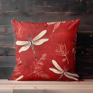 May include: Square red cushion with a dragonfly and floral pattern. The cushion features large cream-coloured dragonflies with black accents and delicate white floral designs. The background is a solid red colour.