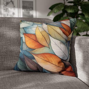 May include: Square throw pillow with a colorful leaf design. The pillow features overlapping leaves in shades of orange, yellow, white, and blue. The pillow is resting on a gray sofa.