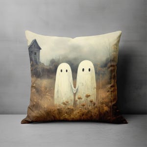 Peut inclure: Coussin carré avec une illustration fantaisiste de deux fantômes blancs se tenant la main dans un champ d'herbes hautes et de fleurs sauvages. Une maison en bois grise et usée est à l'arrière-plan, sous un ciel nuageux. Le coussin a une palette de couleurs neutres.