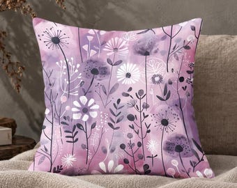 May include: A square throw cushion featuring a watercolour-style background in shades of light purple and pink. The surface is decorated with a repeating pattern of white and dark purple line-art illustrations of various wildflowers and grasses.