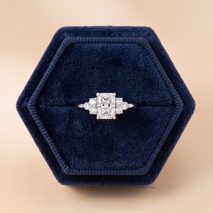 May include: A radiant-cut diamond engagement ring with side stones, set in a silver band, rests in a dark blue velvet ring box. The ring features a large rectangular center stone flanked by smaller stones.