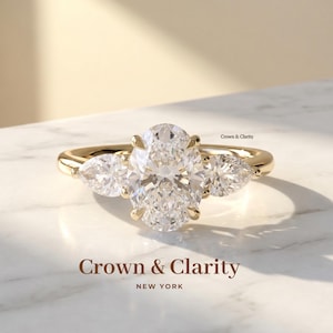 May include: A gold ring featuring a large oval-cut center stone flanked by two pear-cut side stones. The band is polished gold. The text Crown & Clarity New York is visible below the ring, and Crown & Clarity appears near the setting.