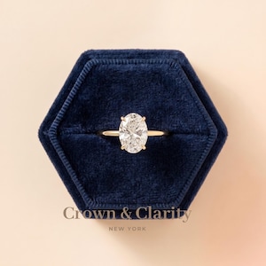 Puede incluir: Un anillo de oro con una gran piedra preciosa transparente de corte ovalado sujeta por cuatro garras. El anillo está centrado en una caja de terciopelo azul oscuro hexagonal. Debajo de la caja, el texto dice Crown & Clarity NEW YORK.