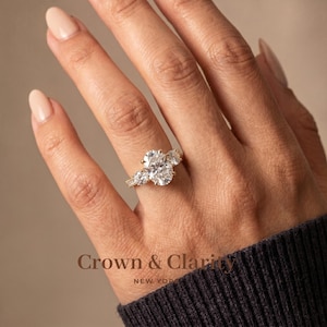 May include: A gold ring featuring a large oval-cut center stone flanked by two smaller round-cut stones on a delicate band. The text Crown & Clarity NEW YORK is visible at the bottom of the image, showcasing a classic three-stone design.