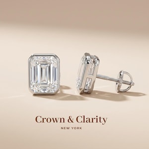 May include: A pair of silver-toned stud earrings featuring emerald-cut clear stones set in a bezel setting. One earring is shown from the front, while the other is shown from the side, revealing the post and screw-back mechanism. Text reads Crown & Clarity NEW YORK.
