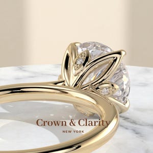 May include: A gold ring featuring a large round brilliant-cut centre stone held by a petal-shaped setting with small accent diamonds. The band is smooth and polished gold. The text Crown & Clarity NEW YORK is visible on the surface.