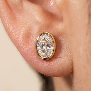 Könnte beinhalten: Nahaufnahme eines goldenen Ohrsteckers mit einem ovalförmigen, klaren Diamanten. Der Diamant ist in einer goldenen Lünettenfassung gefasst. Der Ohrring befindet sich im Ohr einer Person.