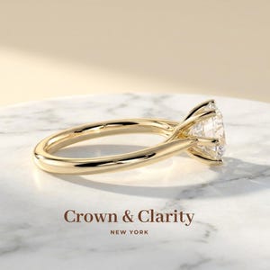 May include: A gold ring featuring a single round brilliant-cut clear gemstone held by a six-prong setting. The band is smooth and polished. The text Crown & Clarity NEW YORK is printed below the ring on the surface.