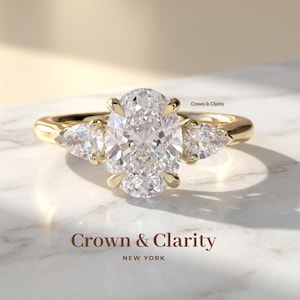 May include: A gold engagement ring with a large oval-cut diamond center stone and two pear-shaped side stones. The ring is set on a gold band. The text "Crown & Clarity NEW YORK" is at the bottom.
