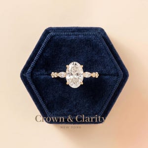 May include: A gold ring featuring a large oval-cut center stone flanked by two smaller pear-shaped stones on each side, set in a dark blue velvet hexagonal box with the text Crown & Clarity NEW YORK printed in gold on the front of the box.