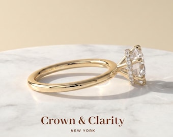 May include: A gold ring featuring a large round centre stone held by a prong setting, with a hidden halo of small pavé diamonds encircling the base of the stone. The text Crown & Clarity NEW YORK is printed below the ring on the surface.