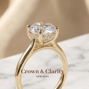 Op de afbeelding: Een gouden ring met een grote ronde briljant geslepen middensteen vastgehouden door vier klauwen, met een verborgen halo van kleinere pavé-gezette stenen onder de hoofdzetting. De tekst Crown & Clarity NEW YORK is zichtbaar op de voorgrond.