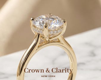 May include: A gold ring featuring a large round brilliant-cut center stone held by four prongs, with a hidden halo of smaller pavé-set stones beneath the main setting. The text Crown & Clarity NEW YORK is visible in the foreground.