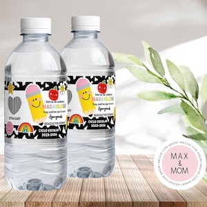 May include: Two clear water bottles with white caps. Each bottle has a label with a black and white pattern, a smiling pencil, a smiling apple, and text in Spanish. The bottles are on a wooden surface.