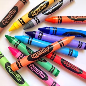 May include: A selection of colourful crayons in shades of brown, yellow, grey, orange, blue, purple, green, pink, and red. Each crayon is labelled with the name "Agustin" in white on a black oval.