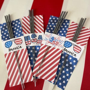 May include: Four packages of sparklers wrapped in red, white, and blue patterned paper. Each package has a different design, including the words "USA", "4th of July", and "America" with sunglasses.