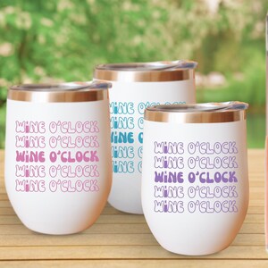 May include: Three white stainless steel wine tumblers with a copper colored rim. The tumblers have the text "Wine O'Clock" repeated in a wavy font in pink, blue, and purple.