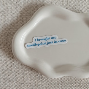 May include: A white, cloud-shaped ceramic tray with a sticker that reads "I brought my needlepoint just in case" in blue text. The tray sits on a textured, neutral-colored surface.