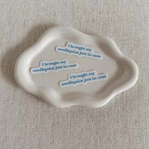 May include: A cream-colored, cloud-shaped ceramic dish holds three stickers. Each sticker has the text "I brought my needlepoint just in case" in a light blue font. The dish sits on a textured, neutral-toned surface.