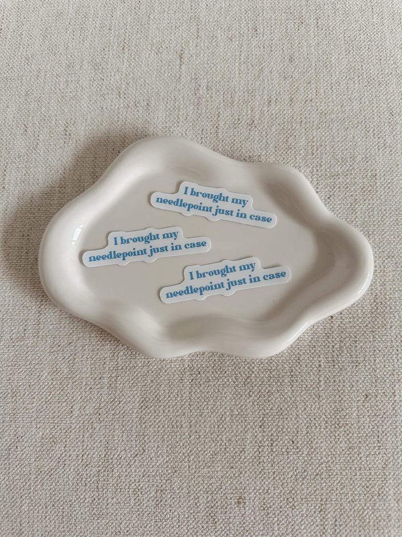 May include: A cream-colored, cloud-shaped ceramic dish holds three stickers. Each sticker has the text "I brought my needlepoint just in case" in a light blue font. The dish sits on a textured, neutral-toned surface.