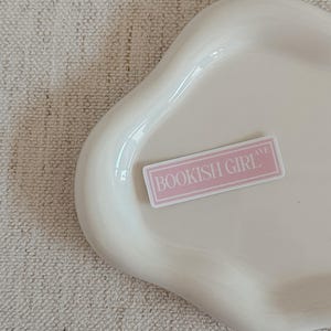 May include: A pale pink sticker with the text "BOOKISH GIRL AVE" in a white rectangular frame rests on a white, organically shaped ceramic tray. The tray is smooth and glossy, set against a textured, off-white background.