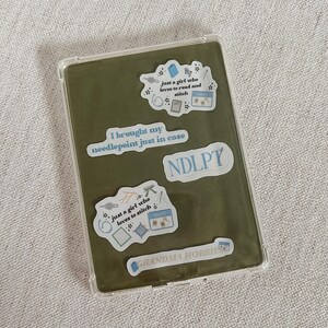 May include: A clear protective case with a green interior, adorned with stickers. The stickers feature text like "just a girl who loves to read and stitch" and "I brought my needlepoint just in case."